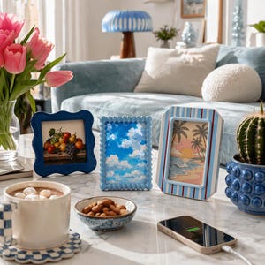 Peut inclure: Trois petits cadres bleus avec des &oelig;uvres d'art de fruits, de nuages et d'une sc&egrave;ne de plage. Une tasse de chocolat chaud, un bol de noix et un t&eacute;l&eacute;phone en charge sont sur une table en marbre. Un vase de tulipes roses et un cactus en pot bleu sont &eacute;galement pr&eacute;sents.
