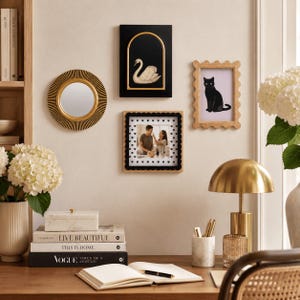 May include: A gallery wall arrangement featuring a round sunburst mirror with a gold center, a black rectangular frame with a gold arch containing a white swan, a scalloped light wood frame with a black cat print, and a square frame with a heart border.