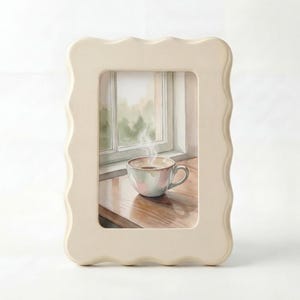 Könnte beinhalten: Ein cremefarbener Bilderrahmen mit gewelltem Rand. Der Rahmen enthält ein Aquarell einer Kaffeetasse auf einer Holzoberfläche vor einem Fenster. Dampf steigt aus der Kaffeetasse auf.