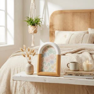 May include: A decorative arch-shaped picture frame with a floral print. The top half is white, and the bottom is natural wood. A small vase with dried flowers, a mug, and a candle sit nearby. A hanging plant and a bed with pillows are in the background.