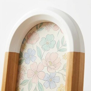May include: An arched picture frame with a white top and wooden base. The frame holds a floral print with pastel pink, blue, and yellow flowers. The frame is a decorative home decor item.