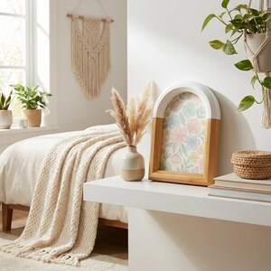 May include: A decorative wooden picture frame with an arched top, featuring a floral print. The frame is white and natural wood. A small vase with dried pampas grass sits nearby, along with a woven basket and a stack of books. The scene is set in a bright, airy room.