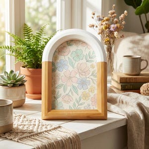 May include: A wooden arch-shaped picture frame with a white top and natural wood base. The frame holds a floral print with pastel pink, blue, and yellow flowers. The frame is displayed on a white windowsill with plants and decor.