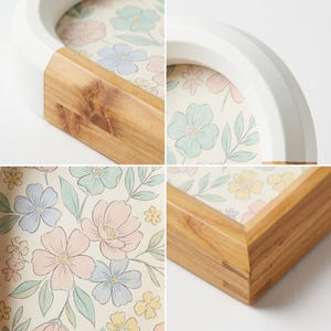 May include: A decorative tray with a floral pattern. The tray has a white, curved rim and a wooden base. The interior features a pastel floral design with pink, blue, and yellow flowers. The tray's wooden frame is visible in the corners.
