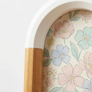 May include: A decorative arched picture frame with a white top and a light wood base. The frame holds a floral print with pink, blue, and yellow flowers on a cream background. The frame's design is modern and minimalist.
