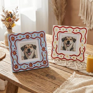 May include: Two square photo frames, each with a picture of a dog. One frame is light blue with red and yellow floral and star patterns. The other is off-white with red, yellow, and blue heart and star designs. Both are on a wooden table.