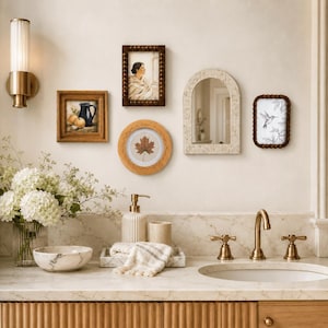 May include: A bathroom vanity featuring a marble countertop, a brass faucet, a white soap dispenser, a marble bowl, and a vase of white flowers. The wall displays a brass wall sconce, two rectangular frames, a round frame, an arched mirror, and a frame.