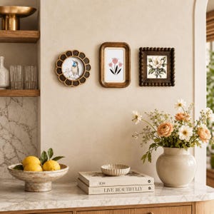 May include: Three wall-mounted picture frames: a circular scalloped frame with a bird illustration, a rectangular rounded frame with a pink flower drawing, and a square beaded frame with a white flower print, displayed above a marble countertop.