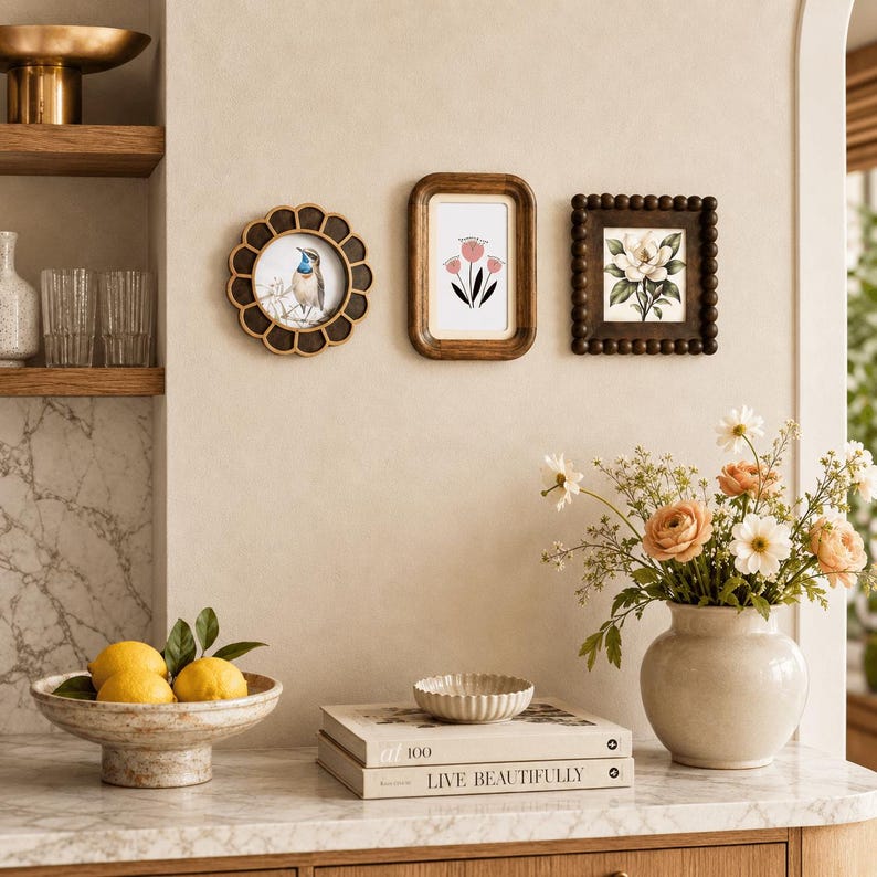 May include: Three wall-mounted picture frames: a circular scalloped frame with a bird illustration, a rectangular rounded frame with a pink flower drawing, and a square beaded frame with a white flower print, displayed above a marble countertop.