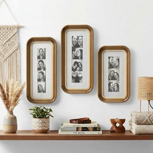 Pode incluir: Três molduras retangulares com cantos arredondados, cada uma com uma moldura dourada e um passe-partout branco. As molduras exibem fotos em preto e branco de pessoas. As molduras são montadas em uma parede branca acima de uma prateleira de madeira.