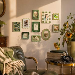 May include: A collection of green decorative picture frames of various shapes and sizes, mounted on a light pink wall. The frames contain images of landscapes, portraits, and botanical designs. A green and white patterned blanket is draped over a leather chair.