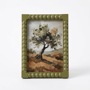May include: A framed landscape painting of a tree in a field. The painting features a tree with green and brown leaves, set against a backdrop of rolling hills. The frame is olive green with a beaded design.