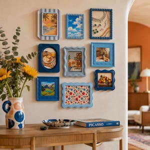 May include: Nine framed pictures in shades of blue are displayed on a cream wall. The frames contain diverse images, such as landscapes and still lifes. A vase of sunflowers and a wooden table with a book and decorative bowl are also visible in the image.
