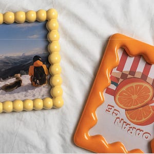 May include: Two decorative picture frames. One is yellow with a bubble design and contains a photo of a person in the snow. The other is orange with a dripping design and features an orange slice graphic with the word "ORANGE".