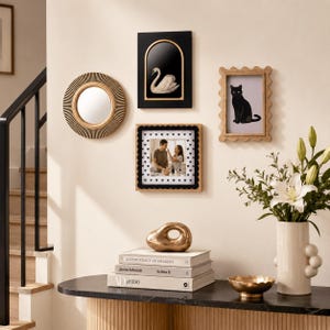 May include: A gallery wall arrangement featuring a round sunburst mirror, a black arched frame with a swan illustration, a light wood scalloped frame with a black cat print, and a square frame with a black scalloped border and a photo of two people.