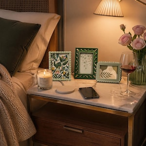 May include: A bedside table with a lit candle, three decorative picture frames, a wireless charger, and a smartphone. A glass of red wine and a vase of pink roses are also present. The frames are green and the candle has a label.