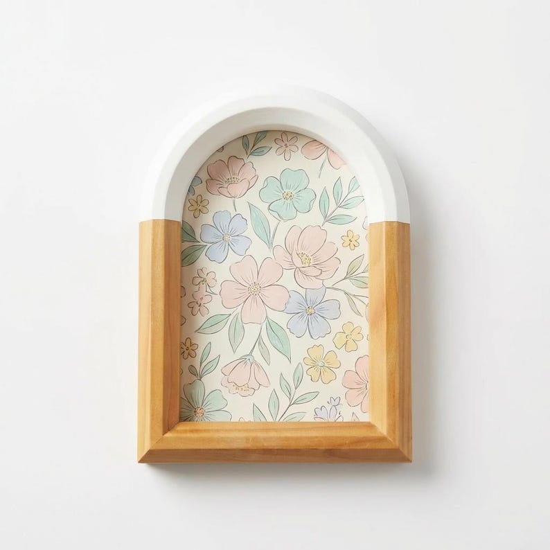 May include: A decorative wooden frame with an arched top. The frame is half-painted white and half natural wood. The interior features a floral print with pastel pink, blue, and yellow flowers on a cream background. The frame is designed for wall display.