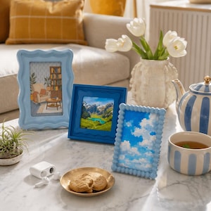 Peut inclure: Trois cadres bleus de diff&eacute;rentes formes sont pos&eacute;s sur une table en marbre. L'un a un bord ondul&eacute;, un autre est carr&eacute; et le troisi&egrave;me a un bord perl&eacute;. Une assiette dor&eacute;e avec deux biscuits, une th&eacute;i&egrave;re blanche et une tasse de th&eacute; sont &eacute;galement sur la table.