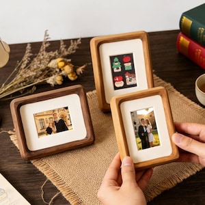 May include: Three wooden photo frames, two with a dark wood finish and one with a lighter wood. The frames contain photos and are displayed on a brown surface. A lamp and books are visible in the background.