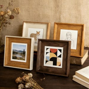 May include: Five square wooden picture frames in shades of brown and white. The frames display different artworks, including a landscape, a geometric design, a portrait, and an animal illustration. One frame has the text "Love is timeless".