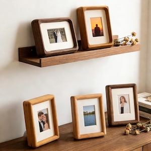 Peut inclure: Plusieurs cadres photo en bois de différentes tailles et teintes, exposant des photographies. Certains cadres sont sur une étagère en bois, d'autres sur une surface en bois. Les cadres ont un passe-partout clair autour des photos.