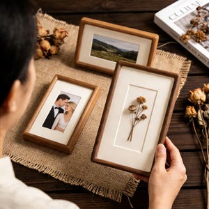 May include: Three wooden picture frames with various images. One frame holds a photo of a couple, another a landscape, and the third features dried flowers. The frames have a light wood finish and white matting.