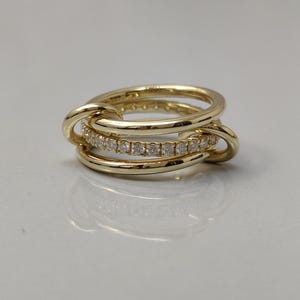 Pode incluir: Um anel dourado com três bandas interligadas. Uma das bandas é adornada com pequenas pedras preciosas transparentes. O anel tem um acabamento polido, realçando o seu design intrincado e artesanato.