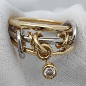 May include: A multi-tone ring featuring gold and silver bands intertwined with a dangling charm. The charm holds a single, round, clear gemstone. The ring's design incorporates a series of interlocking loops and knots.