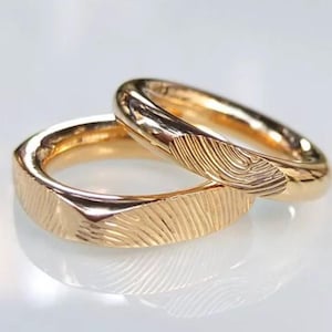 May include: Two polished gold rings featuring a detailed, engraved fingerprint pattern on the outer surface. The bands have a smooth, rounded interior and a textured exterior finish that captures the intricate lines of a human fingerprint.