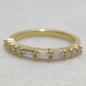 Könnte beinhalten: Ein goldener Bandring mit abwechselnd runden und baguetteförmigen, klaren Edelsteinen. Der Ring hat eine polierte Oberfläche und ist vor einem weichen, neutralen Hintergrund platziert. Der Ring ist ein Schmuckstück.