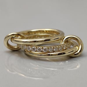 May include: A set of three gold rings interlocked. One ring is encrusted with small, clear gemstones. The rings are made of polished gold and are displayed on a white surface. The rings are designed to be worn together.