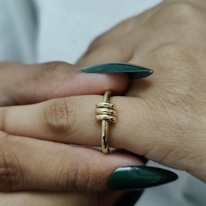 Könnte beinhalten: Ein goldfarbener Ring mit einem einzigartigen Design. Der Ring hat drei gestapelte, abgerundete Elemente in der Mitte, die einen strukturierten Brennpunkt bilden. Das Band ist glatt und schlank. Der Ring wird an einer Hand mit dunkelgrünem Nagellack gezeigt.