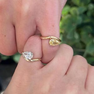 Könnte beinhalten: Zwei goldene Ringe werden gezeigt, einer mit einem birnenförmigen Diamanten und der andere mit einem floralen Design. Die Ringe befinden sich an Fingern, vor einem verschwommenen grünen Hintergrund.