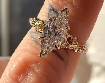 May include: A marquise-cut diamond ring with a gold band. The ring features intricate filigree and small accent diamonds. The main diamond is set in a prong setting, and the band has a floral design.