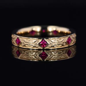 May include: A gold ring featuring a repeating pattern of red square gemstones and detailed scrollwork. The ring is reflected on a black surface, showcasing the intricate design and the vibrant gemstones.