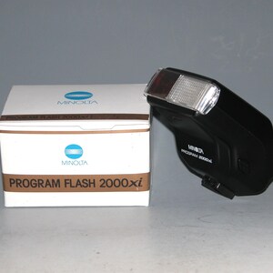 Peut inclure: Un flash d'appareil photo noir Minolta Program Flash 2000xi avec une boîte blanche. La boîte porte le logo Minolta et le texte "PROGRAM FLASH 2000xi". Le flash a un couvercle en plastique transparent et une lentille rouge.