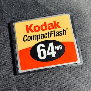 May include: A Kodak CompactFlash memory card with a 64MB capacity. The card has a yellow and orange design, with the Kodak logo in red and the CompactFlash text in black.