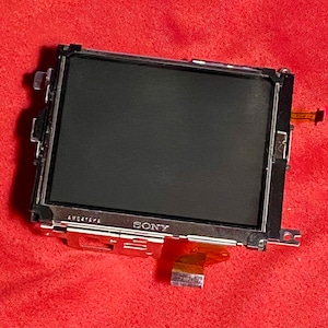 May include: A black LCD screen with a silver frame, featuring the brand name "SONY" on the bottom. The screen has a rectangular shape with a dark display area. It includes a small orange ribbon cable and is set against a red background.