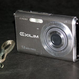 Może przedstawiać: Srebrny aparat cyfrowy Casio Exilim z obiektywem, lampą błyskową i paskiem na nadgarstek. Na aparacie znajduje się napis "EXILIM" i "7.2 MEGA PIXELS". Aparat leży na ciemnej powierzchni.