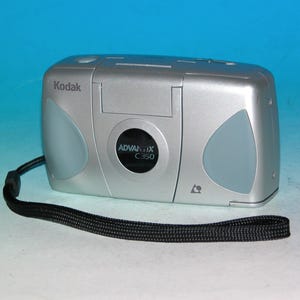 May include: Silver Kodak Advantix C350 camera with a black wrist strap. The camera has a rounded rectangular shape with light blue accents on the sides. The camera has the Kodak logo and Advantix C350 model name.