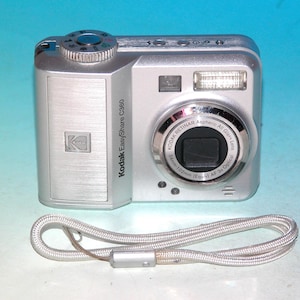 May include: Silver Kodak EasyShare C360 digital camera with a lens, flash, and wrist strap. The camera has the Kodak logo and the text "Kodak EasyShare C360". The lens has the text "KODAK RETINAR Aspheric All Glass Lens".