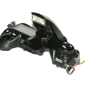 May include: Black Nikon D200 digital SLR camera with the flash open. The camera has a mode dial, ISO and WB settings, and a small LCD screen. The camera is on a white background.