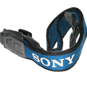 May include: A blue and black camera wrist strap with the word "SONY" in white lettering. The strap is made of woven fabric and has a black plastic connector.