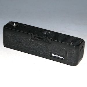 Puede incluir: Un enrollador de película para cámara Kalimar negro con un acabado de imitación de cuero texturizado. El enrollador tiene una pequeña perilla en la parte superior y el nombre de la marca "Kalimar" impreso en el lateral.