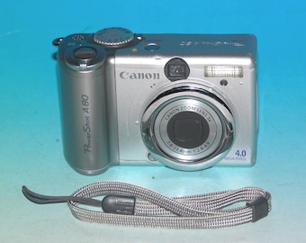 Pode incluir: Uma câmara digital prateada Canon PowerShot A80 com lente de zoom 3x, 4,0 megapíxeis e uma correia de pulso cinzenta. O corpo da câmara apresenta um seletor de modo no topo, um botão de disparo, flash integrado e uma lente com marcações de 7,8-23,4 mm 1:2,8-4,9.