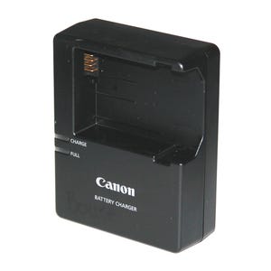 Puede incluir: Un cargador de batería Canon negro. El cargador tiene forma rectangular con una ranura para una batería y las palabras "CHARGE" y "FULL" impresas en el lateral. El logotipo de Canon y "BATTERY CHARGER" están impresos en la parte delantera.