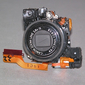 May include: A dismantled Canon camera lens, featuring a zoom lens with the text "CANON ZOOM LENS 3.8x IS 4.6-17.3mm 1:2.8-5.8". The lens is black, with orange and red wiring attached.