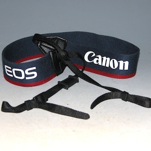May include: A black and red camera strap with the words "EOS" and "Canon" printed in white.