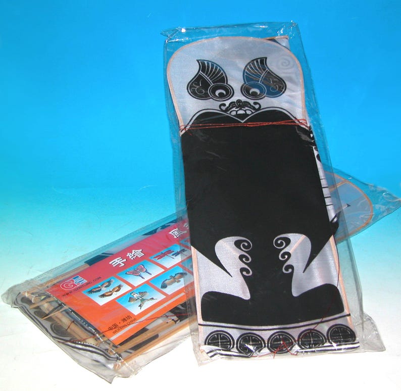 May include: Two kites in plastic packaging. One kite features a black and white design with a face-like pattern. The other package contains a kite with a variety of bird designs. The kites are against a blue background.