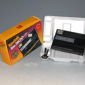Puede incluir: Una cámara Kodak disc 3600 negra en su embalaje original. La caja es amarilla con el logotipo de Kodak y las palabras "Kodak disc 3600 camera" impresas en ella. La cámara tiene un flash incorporado y una correa negra.